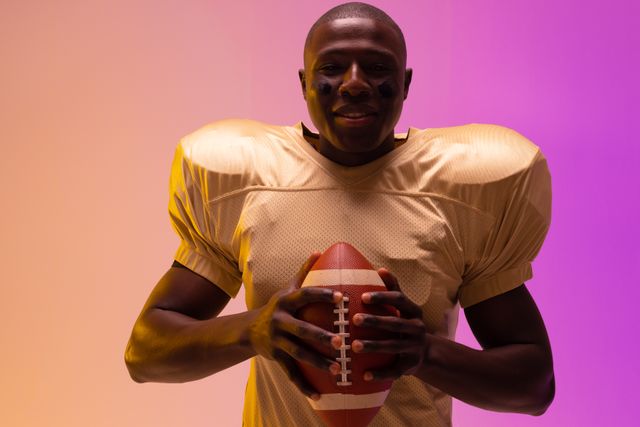 African American football player smiling and holding a football in a vibrant setting with purple and orange lighting. Ideal for use in sports promotions, fitness campaigns, advertisements, and articles about confidence and athleticism.