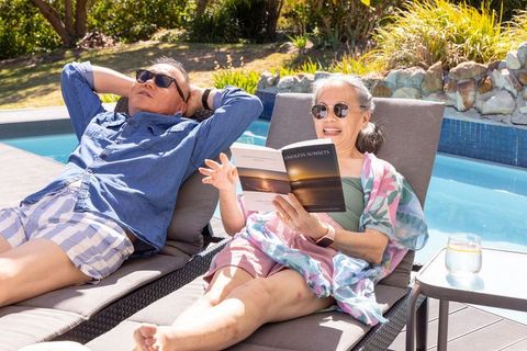 Asian senior couple relaxing on poolside loungers, woman reading 'endless sunsets', enjoying summer