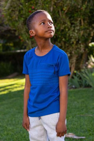 African american boy standing in sunlit backyard wearing blue zip tee and gray sweatpants