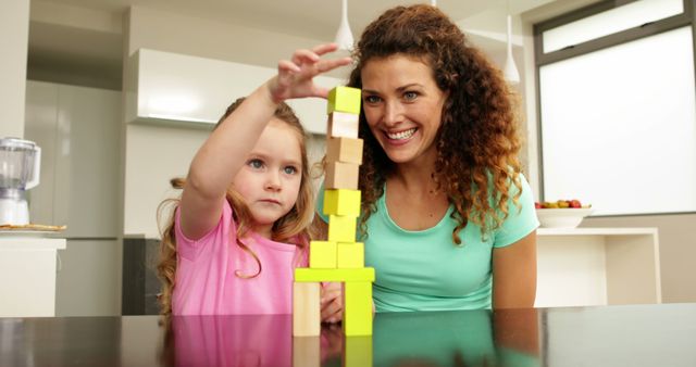 Mother and daughter are engaged in playful activity with building blocks in a modern kitchen. The mother is smiling while the daughter carefully stacks blocks. This scene is ideal for depicting happy family interactions, bonding time, learning through play, and modern home life.
