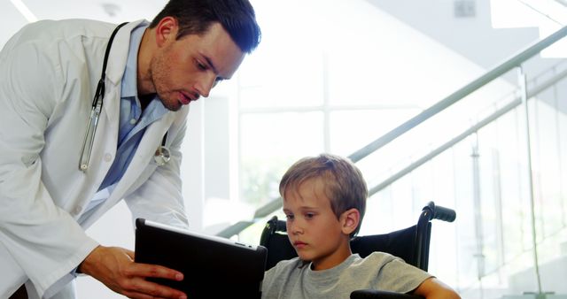 Male doctor using tablet to assist young boy in wheelchair. Clear depiction of healthcare professional interacting with child patient in hospital environment. Effective for health-related articles, medical guides, pediatric consultations, and patient care demonstrations.