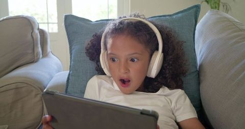 Surprised girl wearing headphones and watching tablet on sofa, reacting to video content