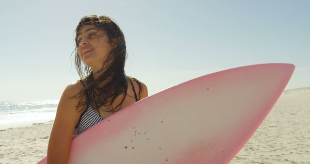 Young woman standing on beach holding pink surfboard. Surfboard indicates active, adventurous mood. Could be used for summer vacation promotions, water sports advertising, women's lifestyle magazines.
