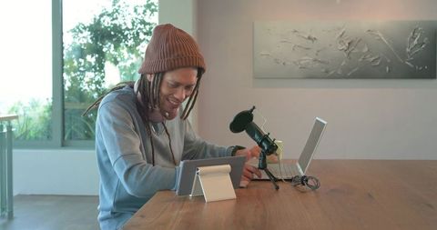 Podcaster recording at home: man with dreadlocks using laptop, tablet, microphone