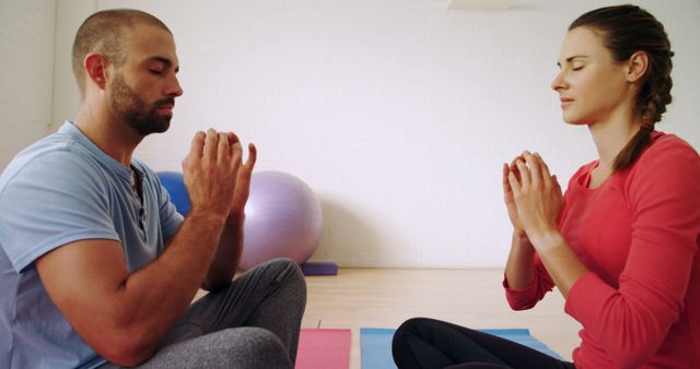 Man and woman sitting cross-legged on yoga mats in peaceful gym while meditating with eyes closed. They appear focused and serene, making this image suitable for promoting yoga classes, wellness retreats, or mindfulness apps.