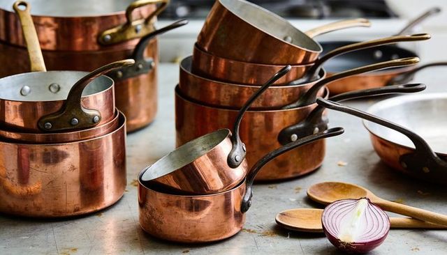 Stacked copper saucepans with wooden spoons and halved red onion on stainless counter