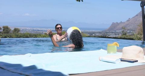 Two women playing beach ball in infinity pool with ocean view and sun hat