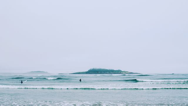 Surfers catching waves on a foggy beach, with distant mist-shrouded islands in the ocean. Ideal for depicting themes of solitude, nature, tranquility, and outdoor adventure. Can be used in travel brochures, serene scene promotions, and water sport-focused content.