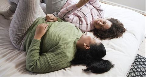 Expecting mother and friend relaxing on bed sharing smartphone and cozy conversation