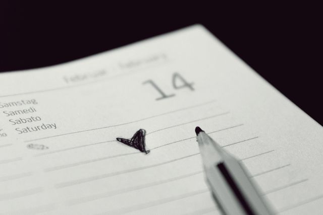 The image shows a close-up of an open diary or calendar, focusing on the date February 14th, often associated with Valentine's Day. A black doodle of a heart marks the day, and a pen lies nearby, suggesting planning or journaling. This image is ideal for content related to Valentine's Day planning, romantic notes, appointments, personal development, and time management.