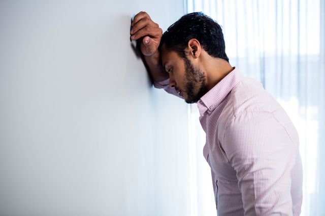 Young businessman in formal attire leaning against wall in office, appearing stressed and thoughtful. Useful for illustrating workplace stress, corporate pressure, mental health in professional settings, and business-related challenges.