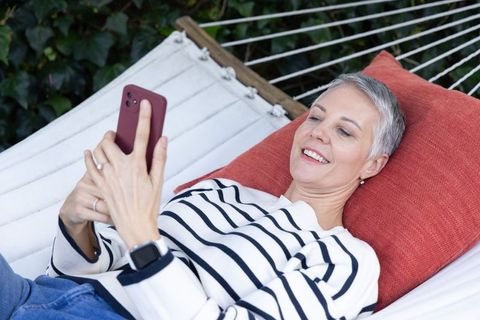 Mature woman relaxing on hammock using smartphone in backyard garden with red cushion