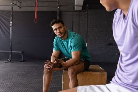 Middle eastern athlete resting on plyo box while talking with workout partner in gym