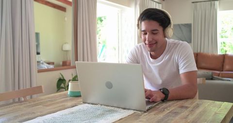 Sunlit african man working remotely on laptop wearing headphones at home dining table