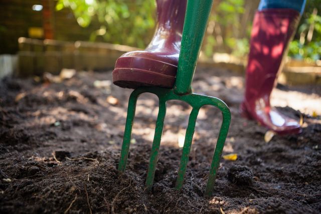 This image depicts a senior woman engaging in gardening activities in her backyard. She is wearing boots and using a garden fork to work the soil. This image can be used for articles or advertisements related to gardening, outdoor activities, senior lifestyle, farming, and agriculture.