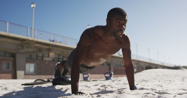 Shirtless man performing push-ups on sandy beach with a bridge and weights in background. Ideal for fitness articles, workout guides, health blogs, and advertisements promoting outdoor exercise and physical strength.