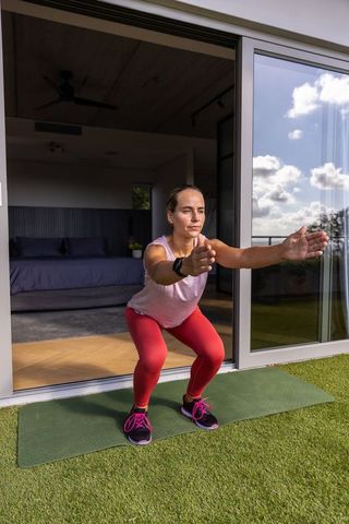 Woman performing bodyweight squat on balcony mat by sliding glass door in red leggings