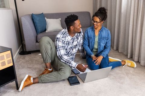 Diverse couple collaborating from home on laptop and tablet while sitting on living room carpet