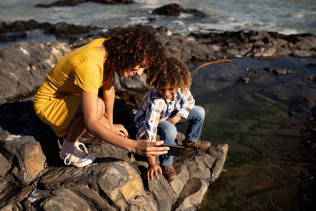 Side view of a biracial woman wearing a yellow dress and her son enjoying time together by the sea, kneeling on rocks on the beach on a sunny day, the owman holding a smartphone and taking a selfie of them