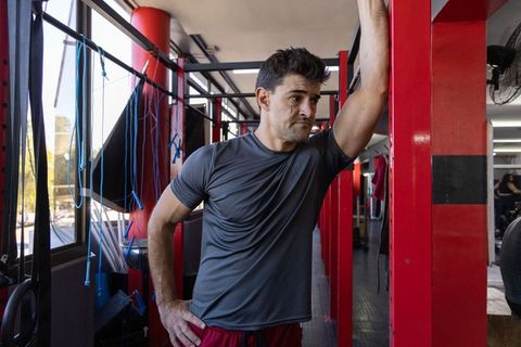 Mid adult man pausing between training rounds leaning on red pillar in sunlit boxing gym