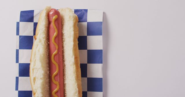 This image of a freshly prepared hot dog with mustard on a blue and white checkered paper background is ideal for promoting food festivals, cookouts, casual dining, and street food vendors. Perfect for use in menus, food blogs, culinary advertisements, and promotional materials for fast food chains.