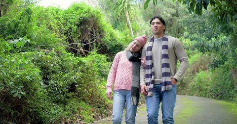 Young couple strolling through lush forest path wearing cozy sweaters and scarves