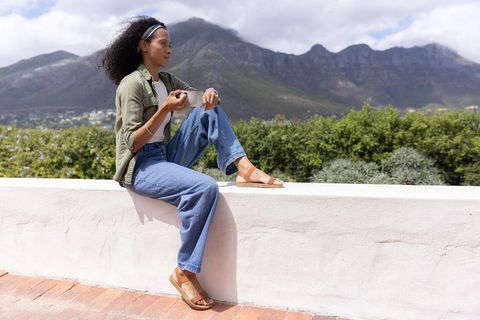 African american woman sitting on white wall holding mug overlooking mountain view and lush garden
