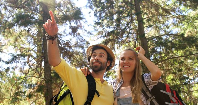 A young Caucasian man and woman are exploring a forest, with the man pointing at something interesting in the distance, with copy space. Their expressions of curiosity and excitement suggest they are on an adventurous hike or nature walk.