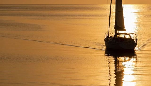 Sailboat silhouette gliding across calm golden sea at sunset with reflected mast and wake
