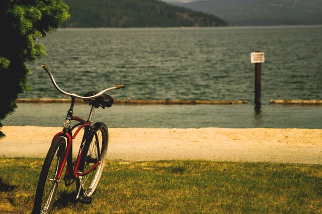 Vintage bicycle resting on grassy lakeshore with tranquil water and forested hills in background. Ideal for portraying outdoor adventure, relaxation, summer vacations, and nostalgic moments.