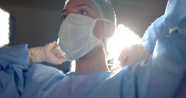 Healthcare worker in scrubs putting on protective gloves and face mask under bright light, emphasizing the importance of safety and preparation in medical fields. Useful for articles about healthcare safety protocols, medical staff preparation, or pandemic responses.