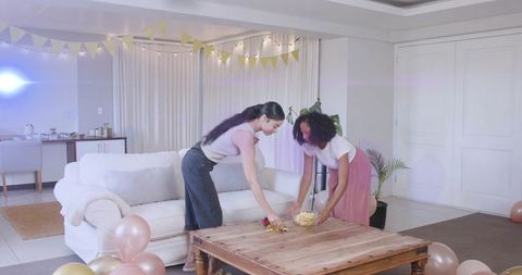 Friends arranging snacks for cozy living room celebration with balloons and bunting