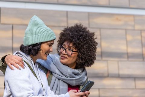 Diverse female friends sharing smartphone and laughter outdoors near urban stone wall
