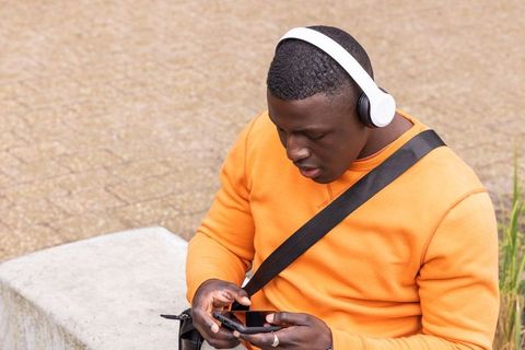 African american man wearing white headphones using smartphone while sitting on urban bench