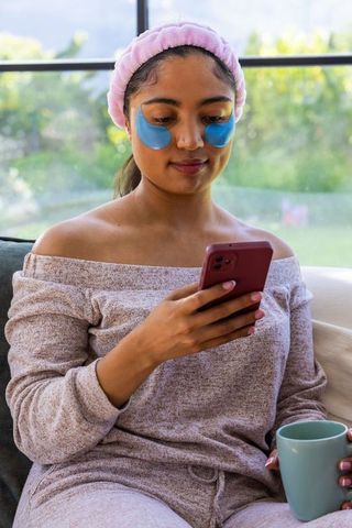 Young woman relaxing with blue under-eye patches using smartphone and holding mug