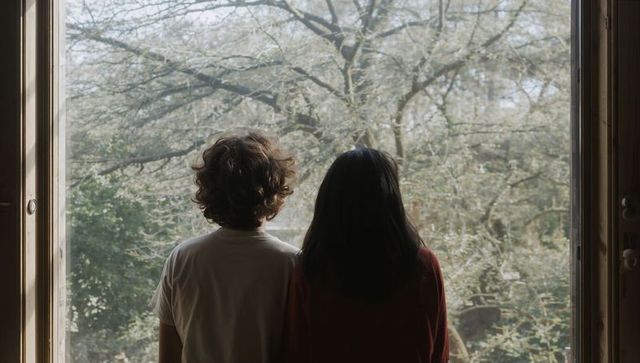 Two teenage siblings standing at large window looking out at trees, contemplative