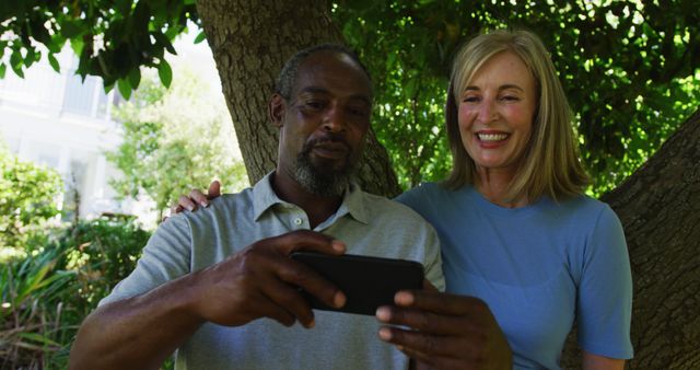 A senior couple is taking a selfie together using a smartphone while standing in a park. They are smiling and appear happy and relaxed. This image can be used for promoting senior lifestyle, technology use among seniors, relationship bonding, diverse couples, or cheerful moments in nature promotions.