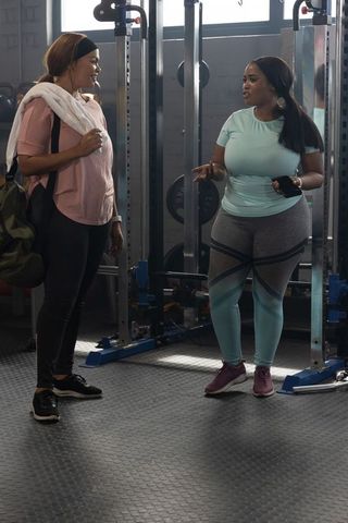 Diverse women chatting at gym squat rack with workout bag and towel