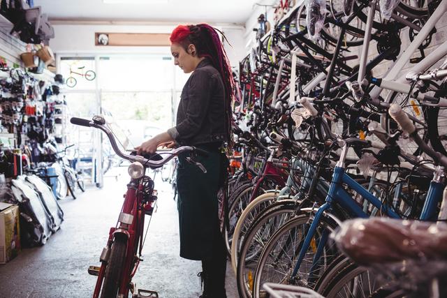 Female mechanic with red dreadlocks working on a bicycle in a shop filled with various bikes and tools. Ideal for use in articles or advertisements related to bicycle repair services, cycling, women in trades, or small business operations.
