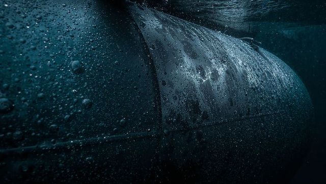 Close-up submerged metal hull showing seam, rivets, corrosion streaks and rising bubbles