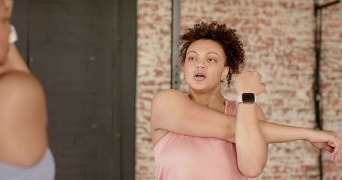 Woman stretching right arm across chest during fitness class wearing smartwatch, pink tank