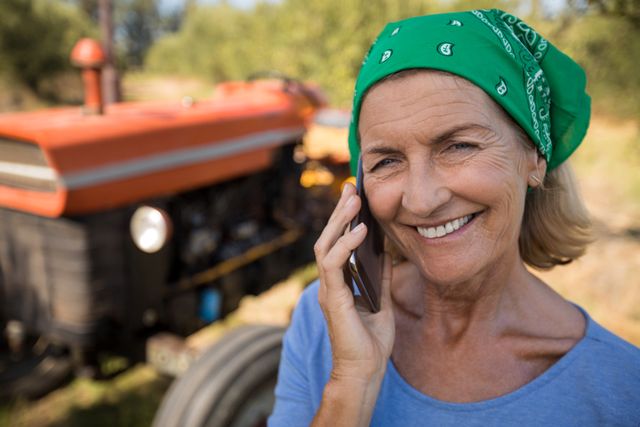 Elderly woman with green bandana smiling while talking on mobile phone in rural setting with tractor in background. Ideal for themes related to senior lifestyle, rural living, agriculture, technology use among seniors, and communication.