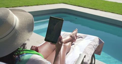 Woman reclining by pool reading tablet wearing sun hat relaxing on lounge chair