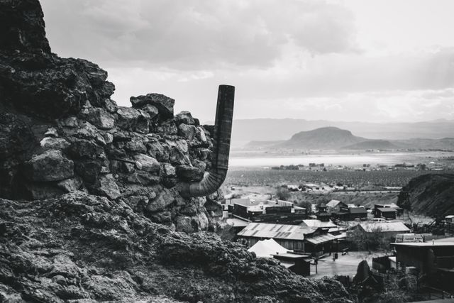 This image shows an abandoned village as seen from a rocky hill, captured in black and white. The landscape includes a cluster of old houses and an industrial pipe protruding from the rock. The mountainous background and the cloudy sky add to the desolate and historical atmosphere. This image is perfect for use in projects about abandoned places, historical sites, and rural landscapes, or to evoke a sense of nostalgia and exploration.