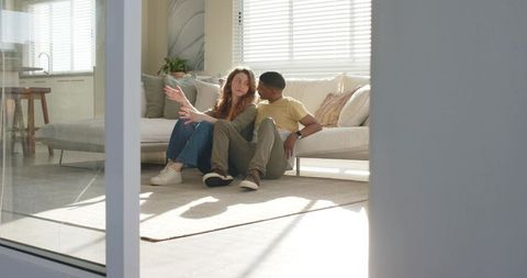 Diverse couple sitting on rug leaning against sofa, sunlit modern interior lifestyle