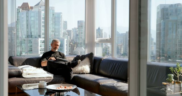 Senior man relaxing on couch in modern apartment with large windows showing city view. Scene depicts comforts of urban living and serenity of retirement. Useful for ads or articles on lifestyle, retirement planning, modern living, and tranquility.