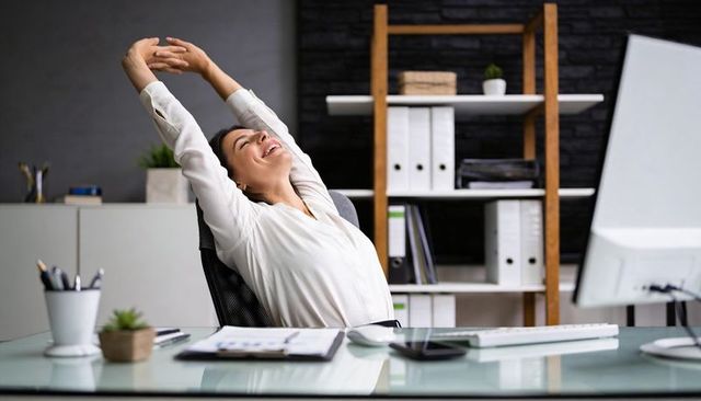 Young woman stretching at desk in modern ergonomic office, smiling with relief, minimalist workspace