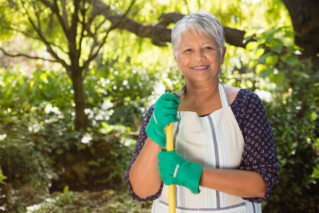 Senior woman standing in a lush garden, holding a gardening tool and smiling. She is wearing green gloves and an apron, enjoying a sunny day outdoors. Ideal for use in articles or advertisements about gardening, senior activities, healthy lifestyles, and outdoor hobbies.