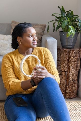 African american woman sitting on rug holding white headphones in cozy sunlit living room