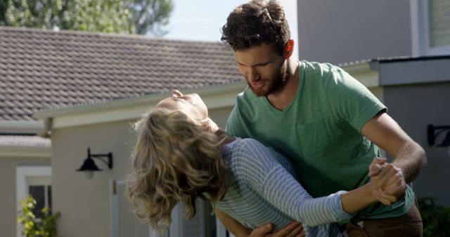 Young couple happily dancing in front of their house during daytime. Ideal for use in advertisements highlighting home life, romance, relationships, or lifestyle blogs focusing on outdoor activities and healthy living.